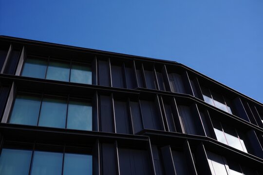 Modern black building facade with large blue-tinted windows under clear blue sky showcasing geometric architectural design and sleek lines - Powered by Adobe
