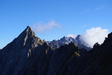 Jagged rocky mountain peaks illuminated by sunlight under a clear blue sky with some clouds enveloping distant peaks