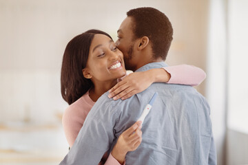 African American man embracing his happy young wife with positive pregnancy test indoors. Millennial black couple expecting baby. Maternity and childbearing concept