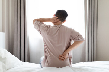 Tired man suffering from back pain after sleeping in bedroom, waking up in morning, sitting on bed...