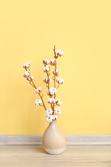 Vase of cotton sprigs on floor near yellow wall