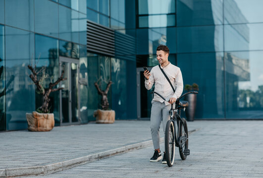 Surprised news and good offer. Shocked cheerful millennial male hipster manager in stylish clothes reads amazing message in phone, stands with bicycle near modern city office on street, outdoor