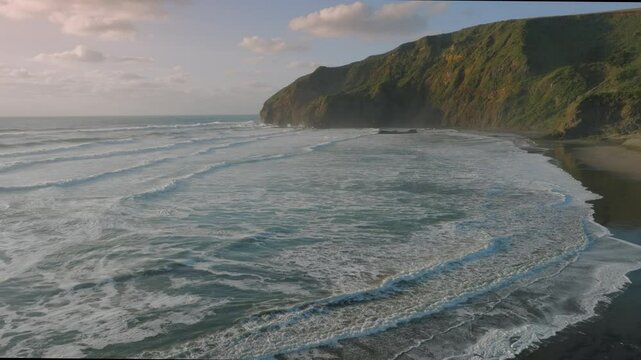 Coastal scene in New Zealand featuring waves crashing on a black sand beach next to a large, green cliff. The ocean is the main focus, with the cliff providing a scenic backdrop. Bethells Beach, NZ