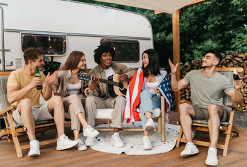 Cheerful interracial friends playing guitar and singing, holding American flag, drinking beer,...