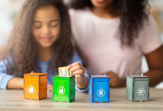 Eco-friendly family activities. Lovely little black girl with her mother playing garbage sorting board game at home, selective focus. Adorable child putting toy trash into tiny bins