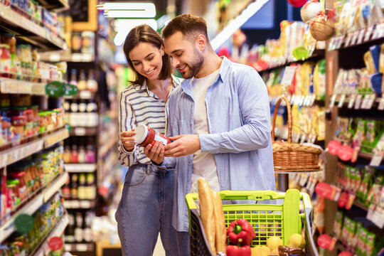 Fototapeta Happy Couple Buying Food In Supermarket, Choosing Products Standing With Trolley Cart Along Aisles And Full Shelves Purchasing Groceries Essentials Together. Smiling Spouses Holding Jar Of Sause