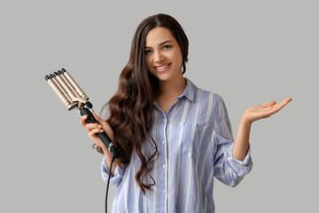 Beautiful young woman with curling iron on grey background
