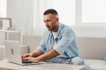 Focused man working on laptop with casual attire in a bright, modern living room, conveying a sense of productivity and creativity in a home office setting.