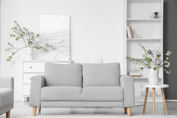 Vase with blooming branches on coffee table near grey sofa in white living room