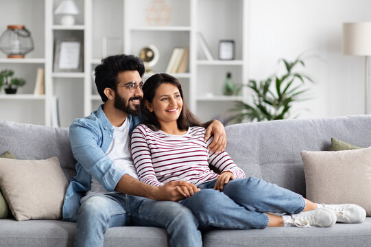 Loving cheerful eastern millennial man and woman in comfy casual outfit cuddling at home, happy sweet couple sitting on couch in living-room, embracing and looking at copy space. Domestic lifestyle