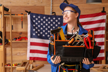Young female carpenter with tool bag and USA flag in workshop. Labor Day celebration