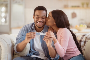 Attractive black woman kissing her husband with positive pregnancy test at home. African American couple happy to find out about their future baby, exchanging caresses indoors