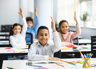 Education, Primary School, Learning and People Concept. Diverse group of smart junior schoolchildren sitting at desks in classroom and raising hands for an answer, studying with pleasure