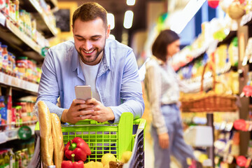 Smiling Male Customer Doing Grocery Shopping Using Smartphone Walking With Cart In Supermarket. Selective Focus. Man Using Groceries Shopping Application On Phone Bying Food In Super Market