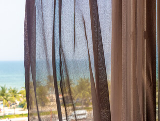 Soft transparent curtains gently blowing beside a window with a tropical view of the sea beach with palm trees