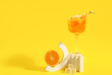 Decorative podiums with glass of Aperol spritz cocktail and half of orange on yellow background