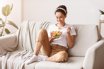 Young and beautiful dark-haired lady sitting on sofa at home, eating colorful fresh vegetable salad and smiling, healthy woman enjoying food full of vitamins, copy space. Food, diet, healthy lifestyle