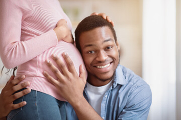 Fototapeta premium Handsome black man listening his wife's pregnant belly at home. Smiling African American future father hugging his expectant girlfriend and touching her tummy, feeling happy indoors