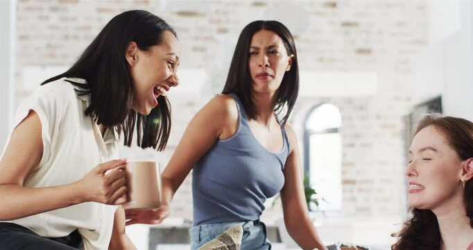 Diverse female friends socializing on loft living room sofa, offering mug and raising drink glass