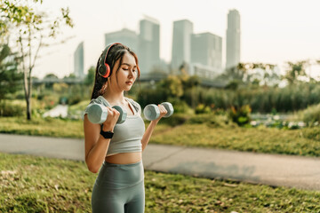 A woman dressed in workout clothes is wearing headphones, listening to music, and lifting dumbbells to exercise her arm muscles.