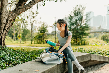 A woman in sportswear sits on a park bench with a duffel bag and water bottle, preparing for an outdoor workout in a green, urban park on a sunny day.