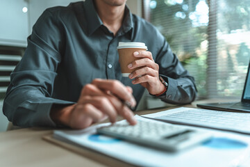 Businessman drink hot coffee. An accountant is seated at a desk in the office, utilizing a calculator to determine balances, profits, currency conversions, and costs.