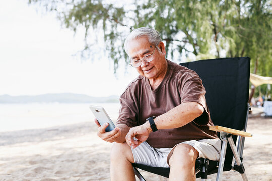 Happy elderly man using smartphone and smartwatch at summer beach camping activity with family outdoors