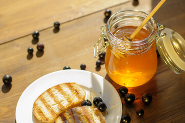 Golden honey in a glass jar sits next to grilled bread on a white plate, with fresh berries scattered around on a rustic wooden table, evoking warmth and flavor.