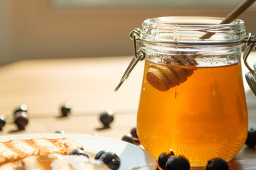 A wooden dipper is seen immersing in a jar of golden honey, casting delightful reflections in the sunlight, surrounded by fresh blueberries on a plate.