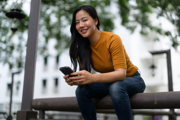 Smiling young asian woman using smartphone while sitting on bench outdoors