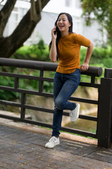 Young Asian woman laughing while talking on mobile phone and leaning on railing in a park
