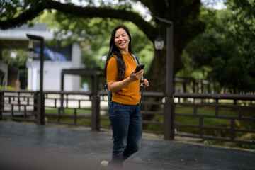 Asian student walking in a park using her smartphone and carrying a backpack