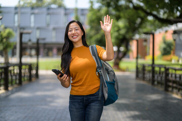 Smiling student waving while walking with backpack and using smartphone in park