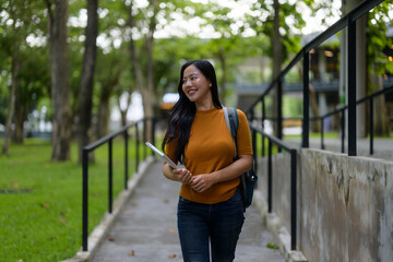Portrait of young happy Asian woman student walking outdoors in university campus holding tablet and backpack