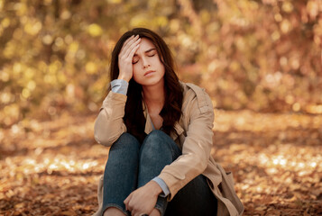 Tired or depressed young woman touching her head at autumn park. Millennial lady suffering from...