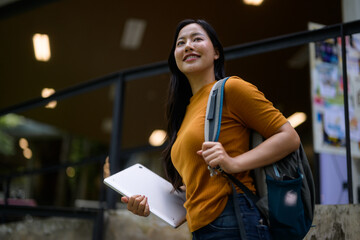 Young Asian student walking up stairs holding tablet and carrying backpack at university