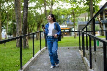 Young woman walking outdoors using mobile phone and holding laptop, enjoying college life in university campus park