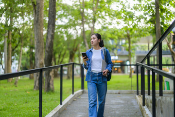 Young woman walking in a green university campus using her smartphone and carrying a laptop