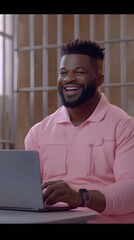 Man wearing a pink top, working on a laptop in a room with bars.