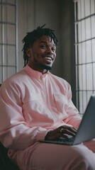 A man wearing a pale pink jumpsuit sits working on a laptop.