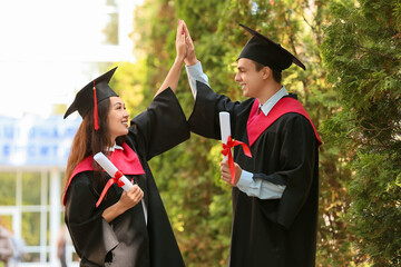 Couple of graduating students with diplomas giving each other high-five outdoors