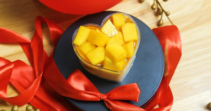 Romantic dessert featuring heart shaped pastry, filled with ripe mango pieces, garnished with red ribbon on dark plate, embodying valentine's day sweetness
