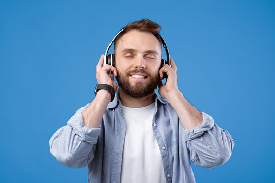 Attractive bearded man in casual outfit listening to music, wearing headphones on blue studio background. Cheerful young guy enjoying popular song with closed eyes