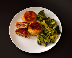 A plate with boiled broccoli and several potato cutlets stuffed with minced meat on a black background.