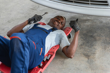 Happy Mechanic Under Car Giving Thumbs Up, Professional Auto Repairman Working in Garage, Sweaty Tradesman Fixing Vehicle on Creeper
