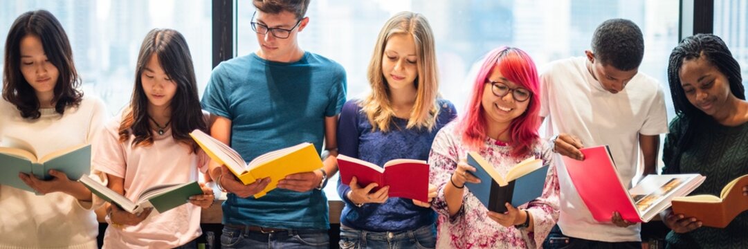 Diverse group of young adults reading books together. Students enjoying reading. Multicultural group of friends with books. Learning and reading in a group. Diverse teenagers reading books at library. - Powered by Adobe