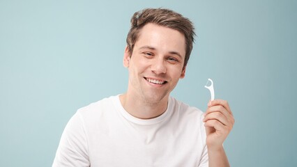Smiling man holding a dental floss pick against a blue background. Dental care, flossing, and oral...