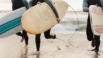 Three surfers carrying surfboards run on the beach. Surfboards in hand, surfers head to the ocean. Beach scene with surfers and surfboards. Extreme sports and adventure travel.