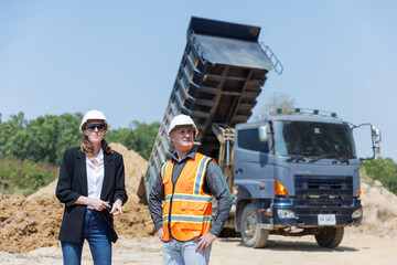 Two construction professionals wearing hard hats and safety gear are discussing near a dump truck...
