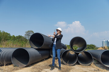 A female construction supervisor in a hard hat and sunglasses uses a walkie-talkie while pointing...